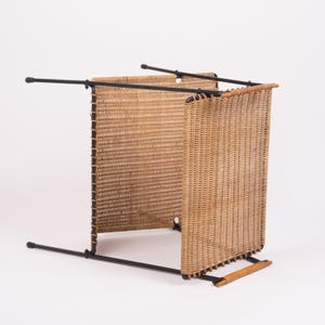 May include: A woven wicker magazine rack with a black metal frame. The rack has a rectangular shape with two woven panels and a handle. The wicker is a light brown color, and the frame is black.