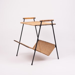 May include: A mid-century modern magazine rack with a woven top and lower shelf. The frame is black metal, and the woven elements are a natural tan color. The rack has two handles at the top and is supported by angled legs.