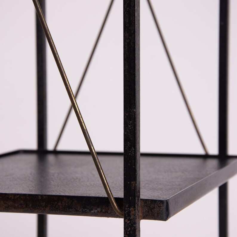 May include: A black metal shelf with three tiers. The shelves are square and the supports are made of thin, dark metal. Diagonal brass-colored metal rods provide additional support. The shelf is against a white background.