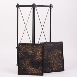 May include: A black metal decorative screen with two square panels. The panels have a textured, speckled design in gold and black. The screen has a tall, vertical frame with intersecting support wires.