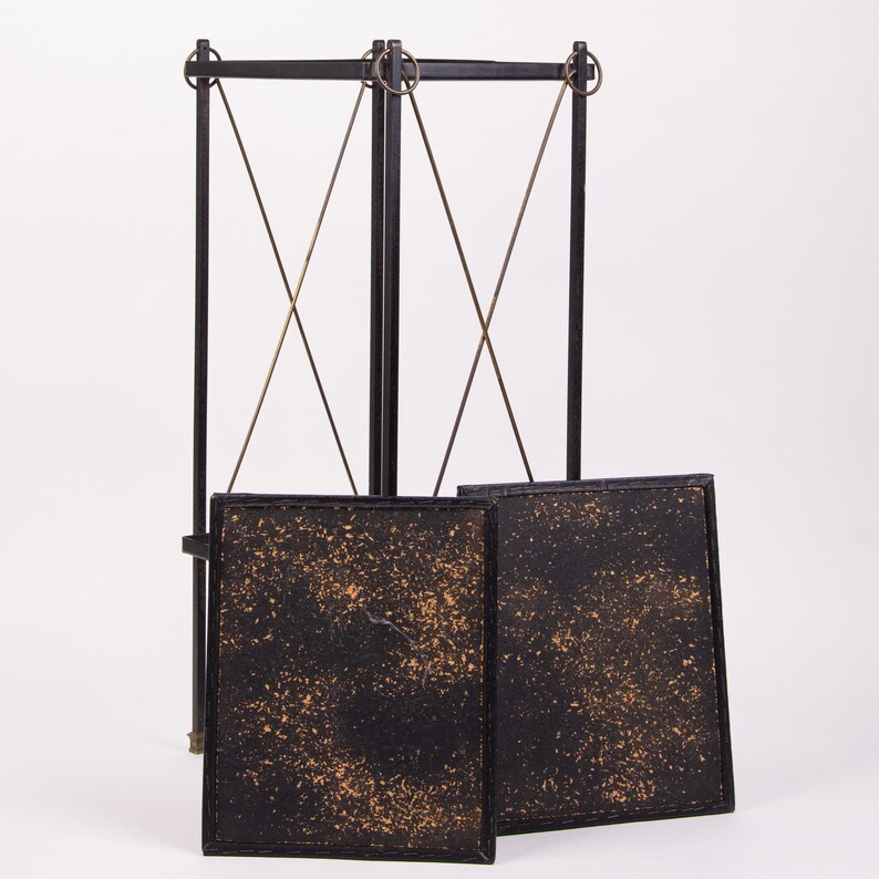May include: A black metal decorative screen with two square panels. The panels have a textured, speckled design in gold and black. The screen has a tall, vertical frame with intersecting support wires.