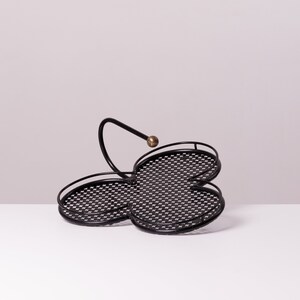 May include: Black metal serving tray with a clover-like design. The tray features three circular sections with a checkered pattern and a curved handle topped with a gold-colored sphere. The tray is designed to hold and display items.