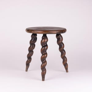 May include: A small, antique wooden stool with a round seat and three intricately carved, spiral-shaped legs. The stool is made of dark brown wood and stands on a white background. The design is reminiscent of a vintage, handcrafted piece.