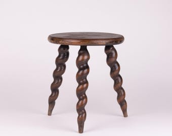May include: A small, antique wooden stool with a round seat and three intricately carved, spiral-shaped legs. The stool is made of dark brown wood and stands on a white background. The design is reminiscent of a vintage, handcrafted piece.