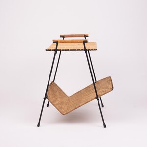 May include: A mid-century modern magazine rack with a woven, light brown basket and tabletop. The rack has a black metal frame and two wooden handles. The design is a stylish blend of form and function, perfect for any living space.