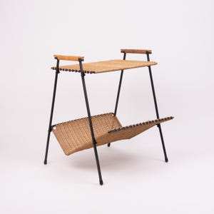 May include: A mid-century modern magazine rack with two woven rattan shelves. The frame is black metal with wooden handles on top. The rack is approximately 24 inches tall and 18 inches wide.