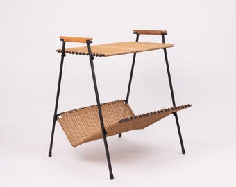 May include: A mid-century modern magazine rack with two woven rattan shelves. The frame is black metal with wooden handles on top. The rack is approximately 24 inches tall and 18 inches wide.