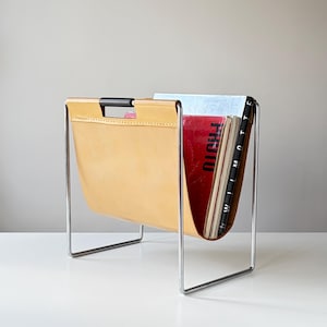 May include: A magazine rack featuring a tan leather sling suspended from a chrome metal frame. The rack includes a black handle integrated into the top of the leather. Several magazines are stored inside, including one with the visible text "PHOTO".
