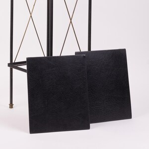 May include: Two black, rectangular, leather-textured panels are displayed in front of a black metal frame with gold-colored accents. The panels are slightly angled, showcasing their textured surface.