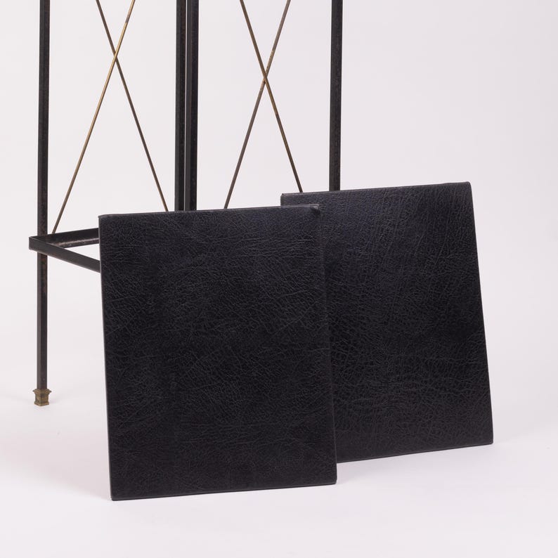May include: Two black, rectangular, leather-textured panels are displayed in front of a black metal frame with gold-colored accents. The panels are slightly angled, showcasing their textured surface.