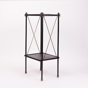 May include: A black metal side table with a square top and a lower shelf. The table has four legs with brass-colored feet and is supported by diagonal metal rods. The top corners are connected by metal rings.