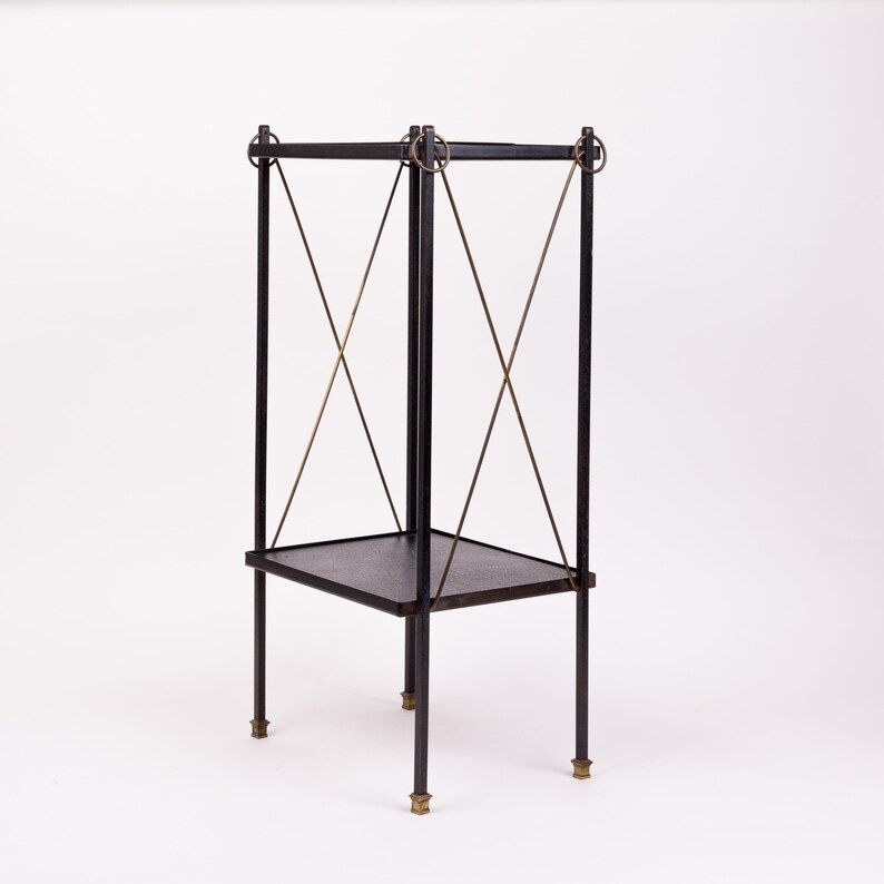 May include: A black metal side table with a square top and a lower shelf. The table has four legs with brass-colored feet and is supported by diagonal metal rods. The top corners are connected by metal rings.
