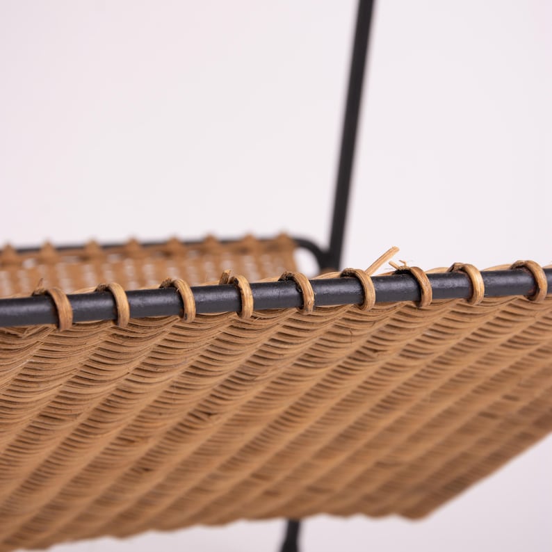 May include: Close-up of a two-tiered shelf with a woven rattan design. The shelves are supported by a black metal frame. The rattan is a light brown color, and the frame is a matte black.