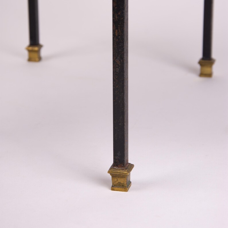 May include: Close-up of a black metal table leg with a square, aged gold-colored foot. The leg has a rectangular shape and a slightly distressed finish. The other legs are visible in the background, also with gold-colored feet.