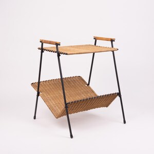 May include: A two-tiered, mid-century modern magazine rack. The rack features a woven rattan shelf and magazine holder, supported by black metal legs. The top shelf has wooden handles. The overall design is a blend of natural and industrial elements.