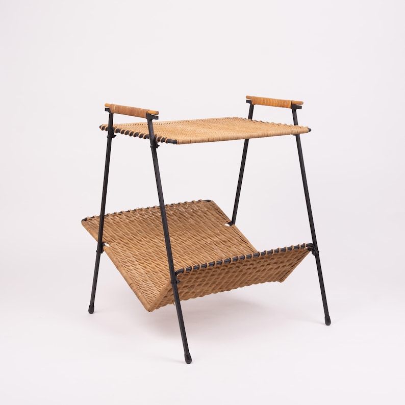 May include: A two-tiered, mid-century modern magazine rack. The rack features a woven rattan shelf and magazine holder, supported by black metal legs. The top shelf has wooden handles. The overall design is a blend of natural and industrial elements.