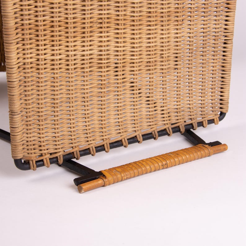 May include: Close-up of a woven wicker chair back with a black metal frame. The chair has a natural tan color and a horizontal handle at the base. The wicker is tightly woven, creating a textured surface.