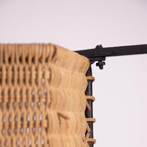 May include: Close-up of a woven wicker panel with a black metal frame. The wicker is a light tan color, and the weave is visible. The metal frame is attached with bolts. The design is likely for a chair or other furniture.