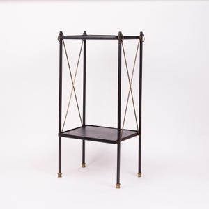 May include: A black metal side table with a rectangular top and a lower shelf. The table has four legs with brass-colored accents at the base and decorative brass-colored crossbars. The top has a dark, textured surface.