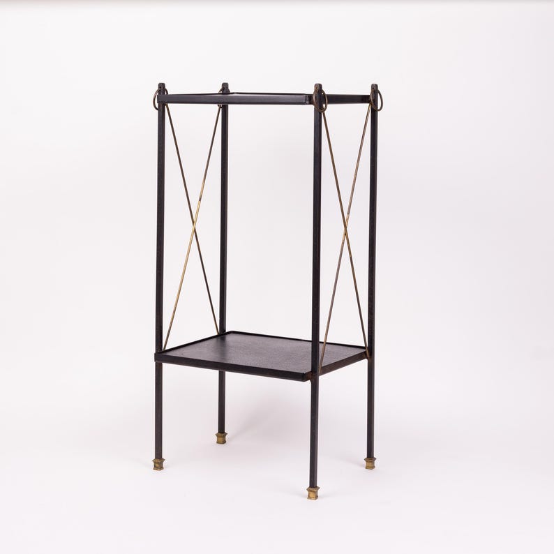 May include: A black metal side table with a rectangular top and a lower shelf. The table has four legs with brass-colored accents at the base and decorative brass-colored crossbars. The top has a dark, textured surface.