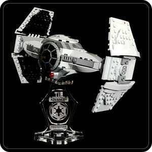 May include: A gray and white TIE Advanced Prototype model, a Star Wars spacecraft, is displayed on a clear stand. The model features detailed wings and a central cockpit. The stand has the text "TIE ADVANCED PROTOTYPE".