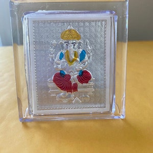 May include: A silver-colored framed religious icon depicting the Hindu deity Ganesha. The icon features Ganesha with a gold crown, blue accents, and red clothing. The frame is clear plastic.