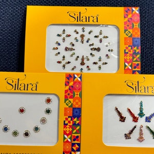 May include: Three yellow packages with the word "Silara" printed on them. Each package contains a sheet of colorful rhinestones for body art.