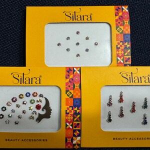 May include: Three yellow boxes with the word "Silara" printed on them. Each box contains a sheet of colorful adhesive gems for decorating the face or body. The gems are arranged in different patterns on each sheet. The boxes are labeled "Beauty Accessories".