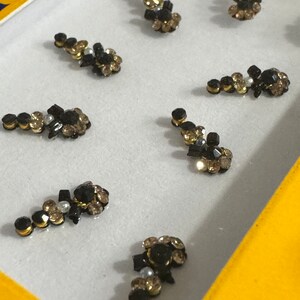 May include: A set of eight black and gold rhinestones arranged in different patterns. The rhinestones are designed to be applied to the face as a decorative accent.