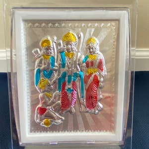 May include: A framed silver religious artwork featuring three figures with gold and colorful accents. The figures are set against a silver background with a sunburst design. The artwork is encased in a clear plastic frame.