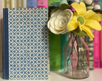 May include: A hardcover book with a blue and white floral pattern next to a small glass vase with felt flowers. The flowers are yellow, white, and green. Books of various colors are in the background.