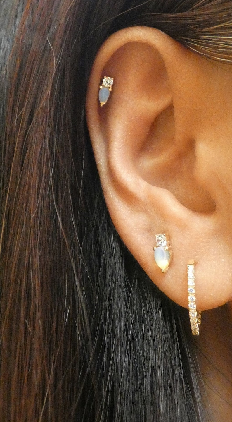 May include: Close-up of an ear with three gold earrings. The upper ear has a stud with a clear stone and a teardrop-shaped, light blue stone. The middle ear has a similar stud. A small hoop earring with clear stones is in the lobe.