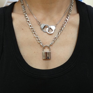 May include: A silver-toned necklace set featuring three separate pieces. The top necklace has a small chain with a pair of handcuffs. The middle necklace has a thick chain with a padlock pendant. The bottom necklace has a thick chain.