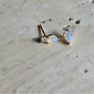 May include: A pair of gold-coloured stud earrings. Each earring has a pear-shaped, opalescent stone and a small, square-cut clear stone. The earrings are displayed on a light-coloured, textured surface.