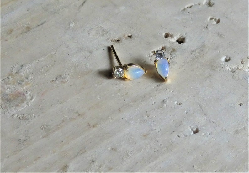 May include: A pair of gold-coloured stud earrings. Each earring has a pear-shaped, opalescent stone and a small, square-cut clear stone. The earrings are displayed on a light-coloured, textured surface.