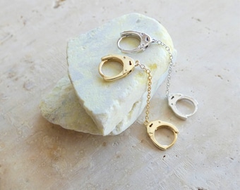 May include: Two pairs of silver and gold handcuff earrings with a chain connecting the two pieces. The earrings are displayed on a white rock.