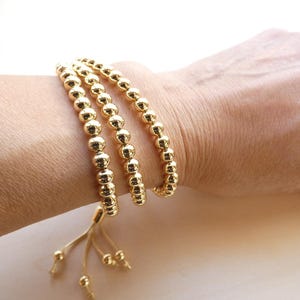 May include: Gold beaded bracelet wrapped around a wrist. The bracelet features multiple strands of small, round, gold beads. The bracelet is secured with a gold tassel and small gold beads at the end. The bracelet is a jewelry item.