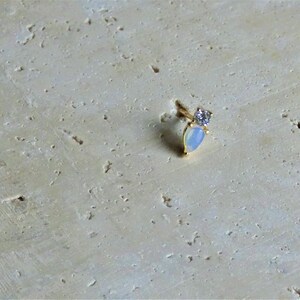 May include: A small, gold earring featuring a pear-shaped, opalescent stone and a small, clear gemstone. The earring is set against a textured, light beige background.