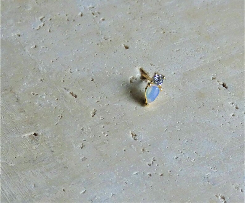 May include: A small, gold earring featuring a pear-shaped, opalescent stone and a small, clear gemstone. The earring is set against a textured, light beige background.