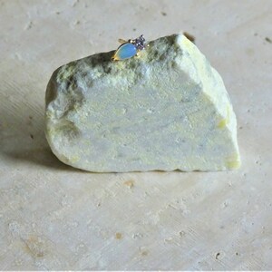 May include: A gold ring featuring a pear-shaped, pale blue opal and a cluster of small, clear stones. The ring is displayed on a light green and yellow rock, against a neutral backdrop.