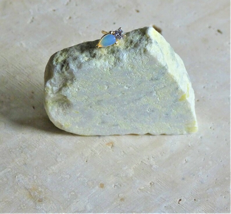 May include: A gold ring featuring a pear-shaped, pale blue opal and a cluster of small, clear stones. The ring is displayed on a light green and yellow rock, against a neutral backdrop.