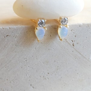May include: A pair of gold stud earrings. Each earring has a clear, round gemstone above a pear-shaped, opalescent stone. The earrings are displayed on a textured, light-coloured surface.
