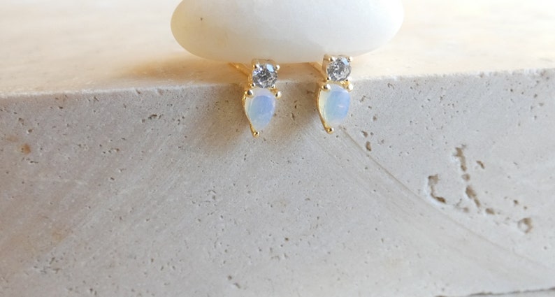May include: A pair of gold stud earrings. Each earring has a clear, round gemstone above a pear-shaped, opalescent stone. The earrings are displayed on a textured, light-coloured surface.