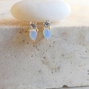 May include: Gold-coloured stud earrings featuring a pear-shaped, opalescent stone and a small, square, clear stone. The earrings are presented on a textured, light-coloured surface.