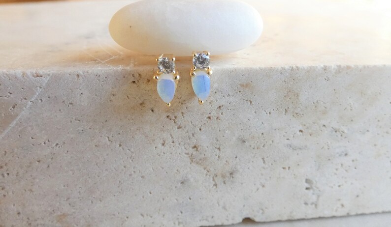 May include: Gold-coloured stud earrings featuring a pear-shaped, opalescent stone and a small, square, clear stone. The earrings are presented on a textured, light-coloured surface.