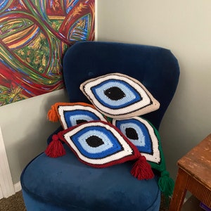 以下が含まれることがあります： 青、赤、緑、ベージュなど、さまざまな色のクロシェで編まれた邪眼のクッションが4つ、青いベルベットの肘掛け椅子に置かれています。クッションはダイヤモンド型で、中央に大きな青い目があります。