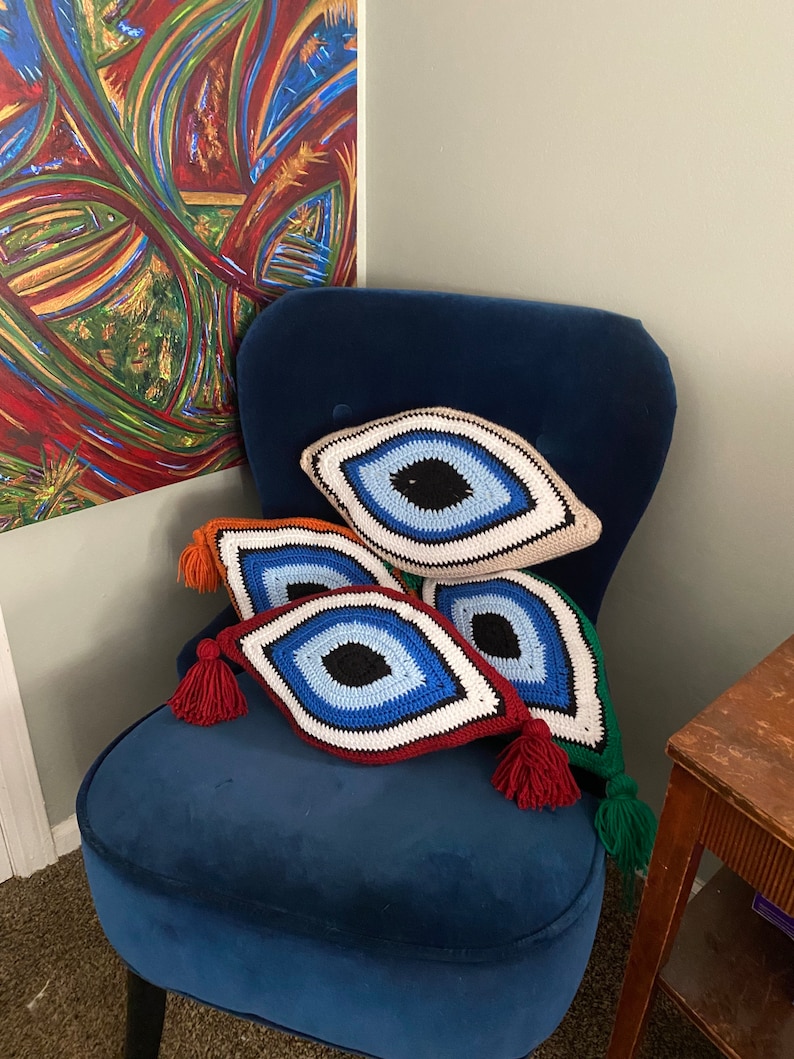 以下が含まれることがあります： 青、赤、緑、ベージュなど、さまざまな色のクロシェで編まれた邪眼のクッションが4つ、青いベルベットの肘掛け椅子に置かれています。クッションはダイヤモンド型で、中央に大きな青い目があります。