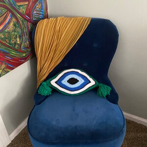 以下が含まれることがあります： 緑色の房飾り付きの、青いベルベットの肘掛け椅子に、クロシェで編まれた青い目のクッションが置かれています。クッションは青、白、黒で、中央に青い目があります。