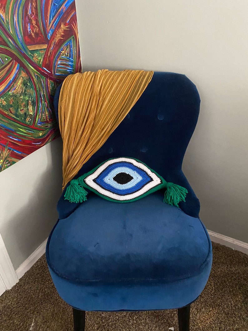以下が含まれることがあります： 緑色の房飾り付きの、青いベルベットの肘掛け椅子に、クロシェで編まれた青い目のクッションが置かれています。クッションは青、白、黒で、中央に青い目があります。