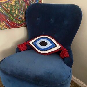 以下が含まれることがあります： 赤い房飾りがついた、青いベルベットの肘掛け椅子に、クロシェで編まれた邪眼のクッションが置かれています。クッションは白、赤、青、黒です。